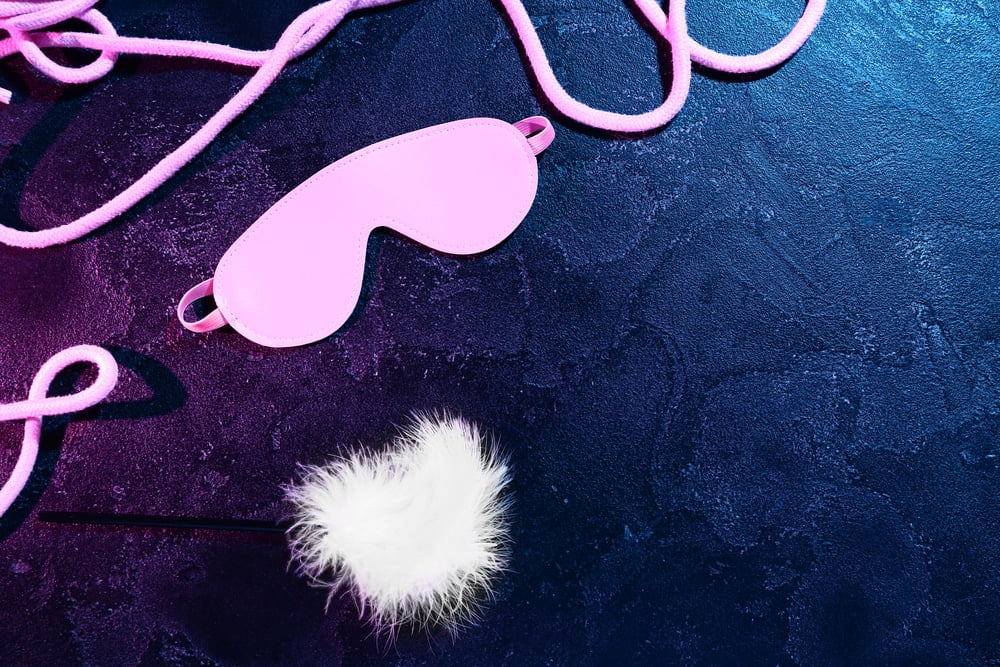 Pink blindfold, soft white feather, and rope arranged on dark textured background under neon light
