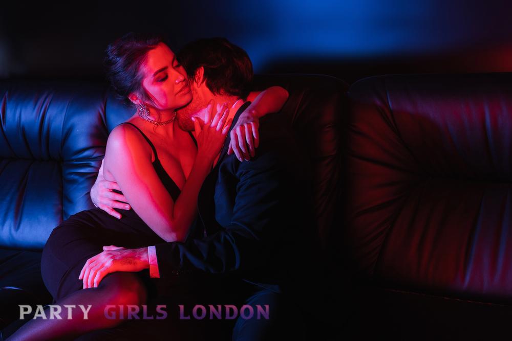 Elegant couple sharing an intimate moment on a leather sofa under red and blue lights, symbolising desire and connection in the refined world of London escorts