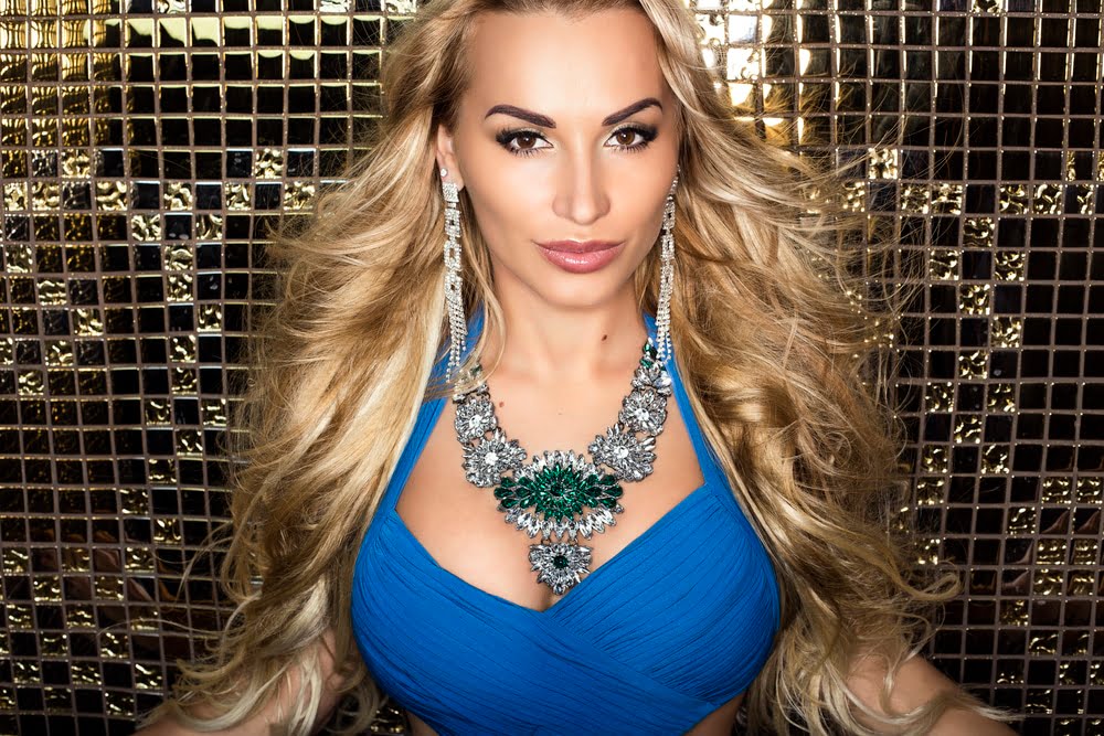 Strapon Fun with a Fulham Escort in a blue dress and statement jewellery posing against a gold mosaic background