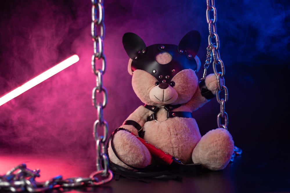 Teddy bear wearing playful leather accessories sitting among metal chains in neon pink and blue light