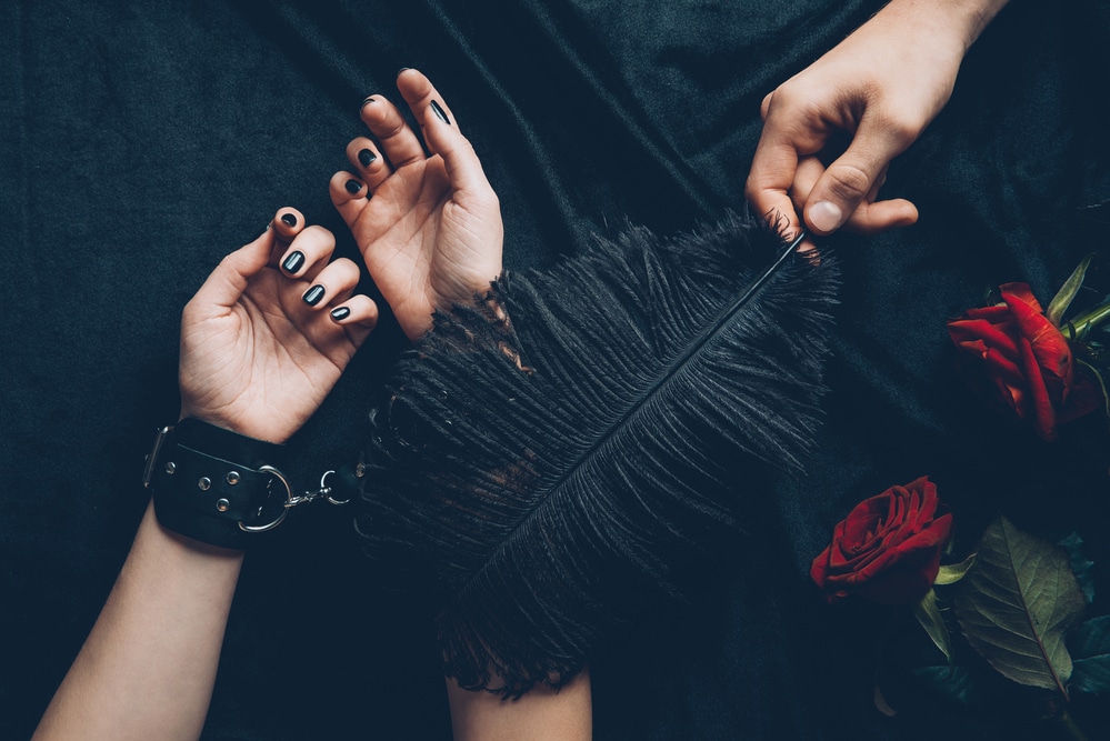 Close-up of hands on dark fabric with a feather and roses, symbolising playful intimacy and trust