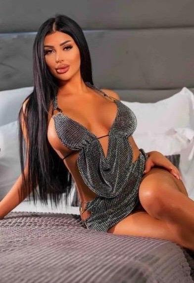 Natalie reclines on a bed wearing a shimmering dress. Her long black hair cascades over her shoulders.